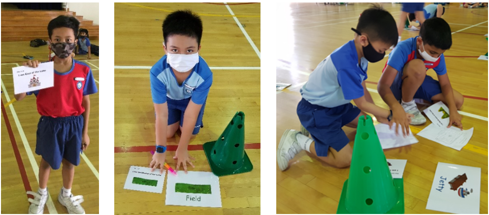 Three masked boys in blue uniforms do a floor activity with green cones and word cards on a gym floor.
